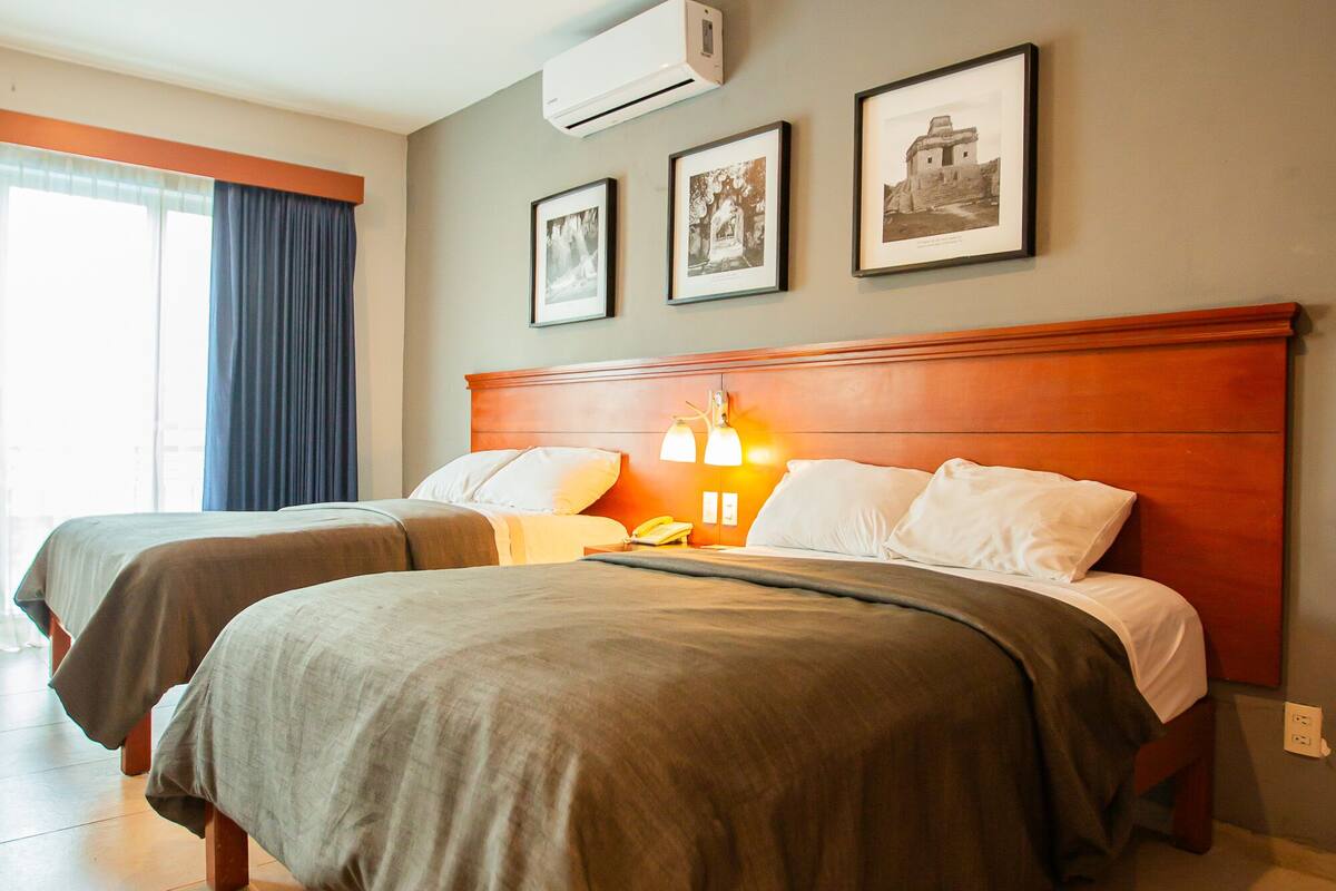 A hotel room with two beds, a wooden headboard, a window with curtains, and framed pictures on the wall.