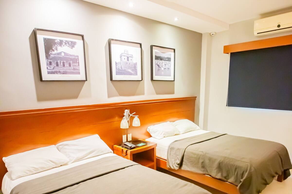 A hotel room with two single beds, a wooden headboard, a nightstand with a lamp, and framed pictures on the wall.