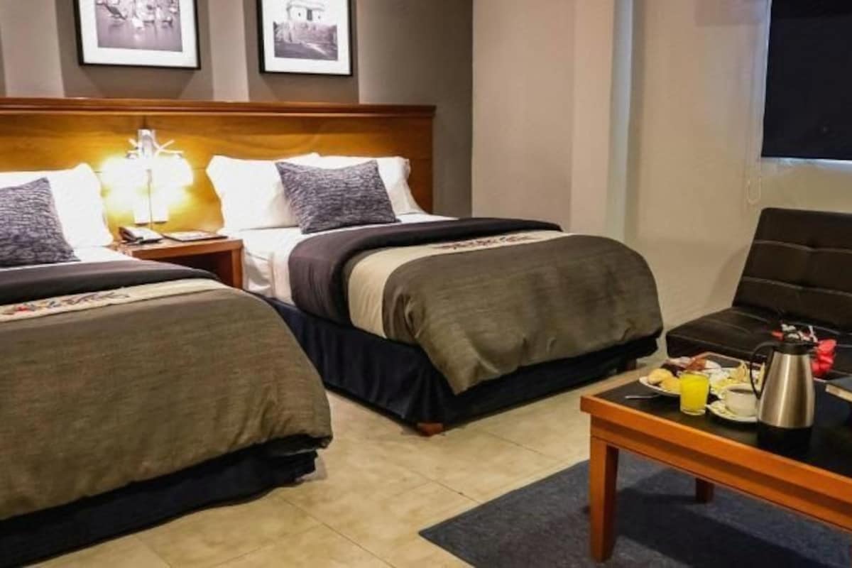 A hotel room with two beds, a sofa, a coffee table with breakfast items, and a TV.
