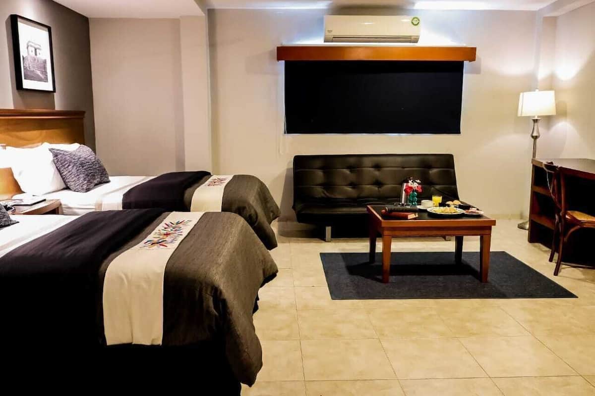 A hotel room with two beds, a sofa, a small table with breakfast items, an air conditioner, and a TV mounted on the wall.
