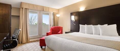 A hotel room with a large bed, a red armchair, a desk with a chair, and a view of the outdoors through a sliding door.