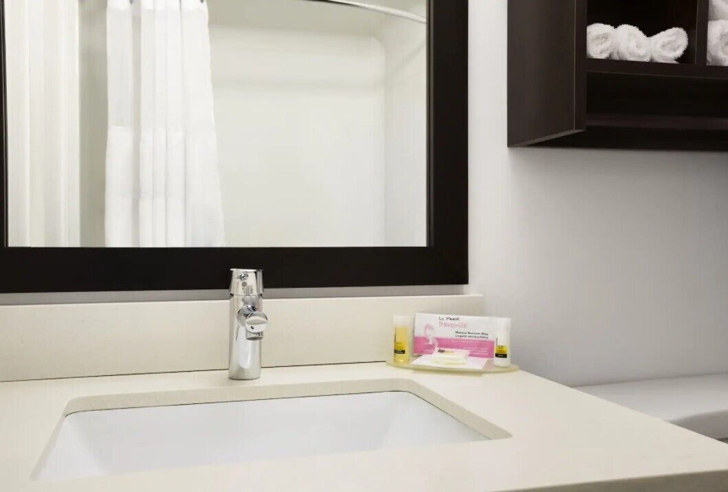 Un baño con lavamanos blanco, un espejo con marco negro y una estantería con toallas.