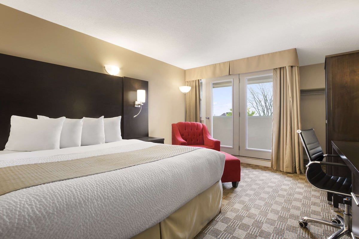 A hotel room with a large bed, a red armchair, a desk with a chair, and a view of the outdoors through a sliding door.