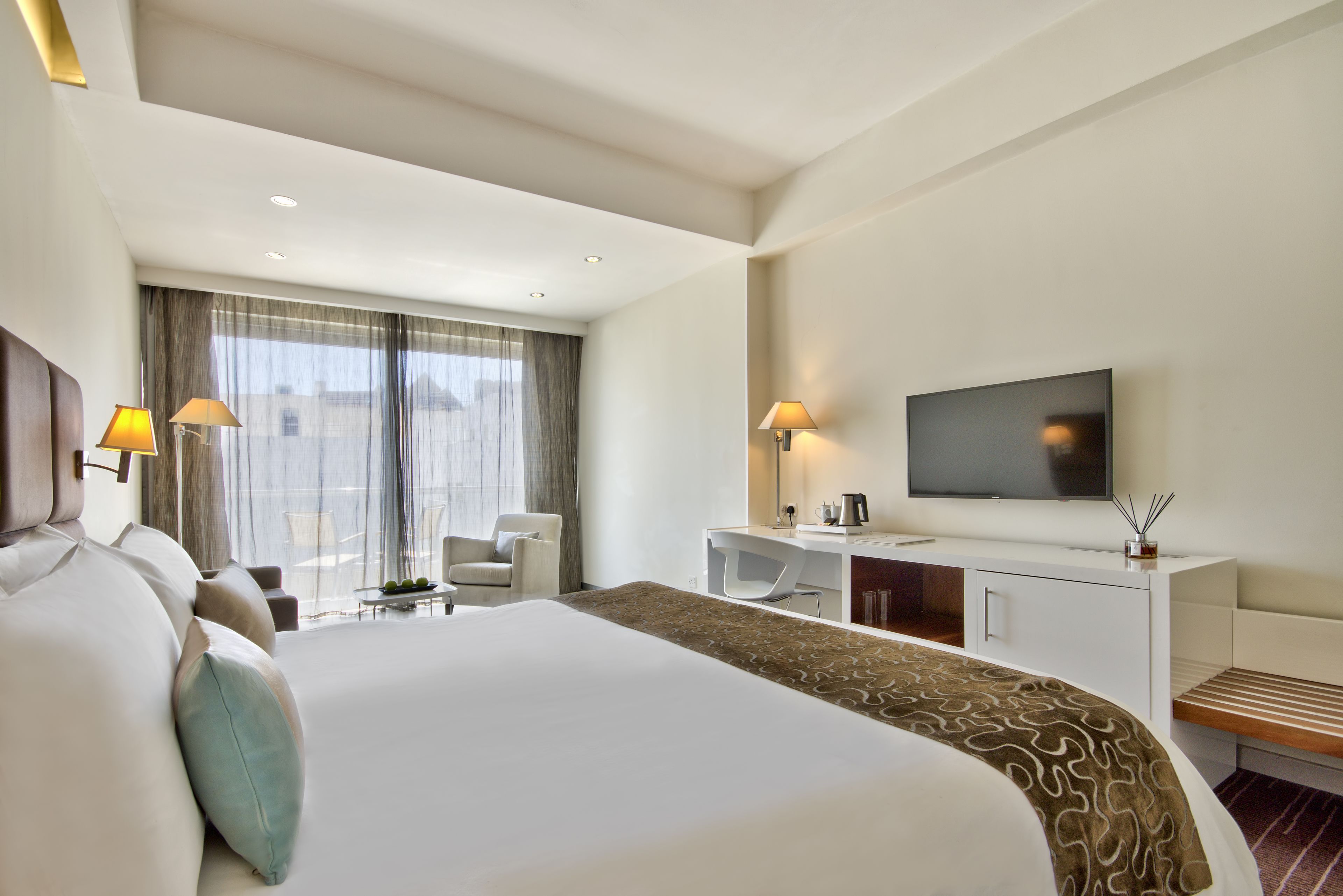 A modern hotel room with a large bed, a TV, a desk, and a seating area with a view of the city.