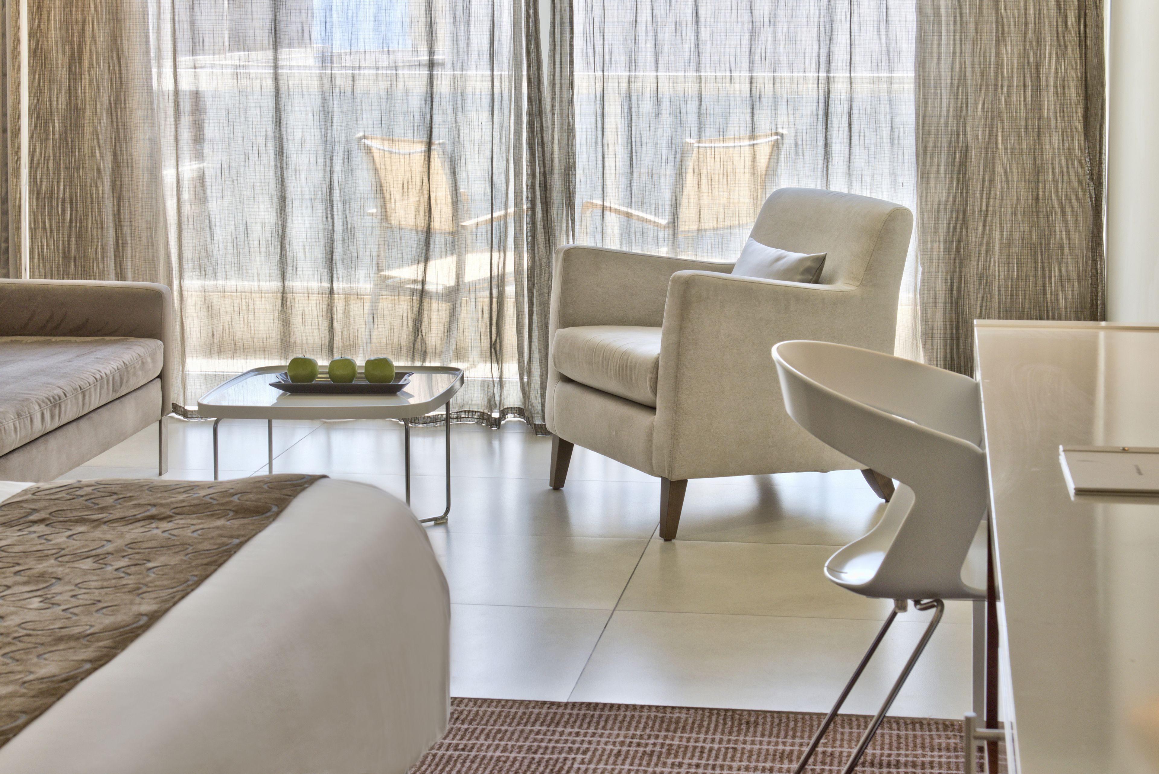A modern hotel room with a bed, a sofa, a chair, a small table with apples, and sheer curtains.