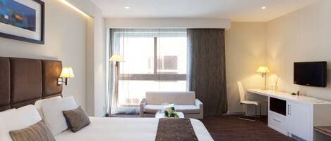 Habitación de hotel con una cama grande, un escritorio, una silla, un televisor y un ventanal con cortinas.