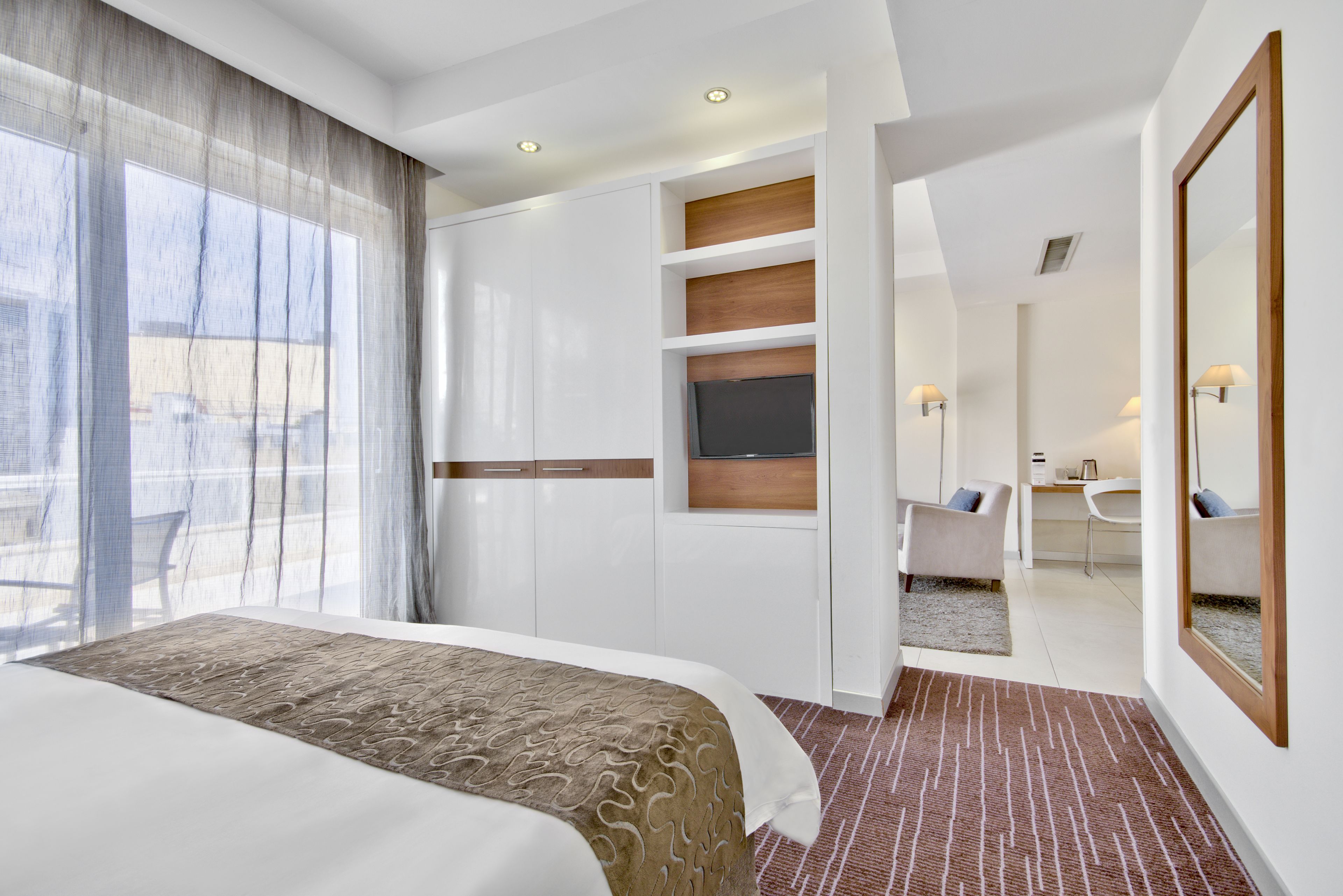 Een moderne hotelkamer met een groot bed, een bureau en een stoel. Er is een raam met gordijnen, een flatscreen televisie en een spiegel aan de muur.