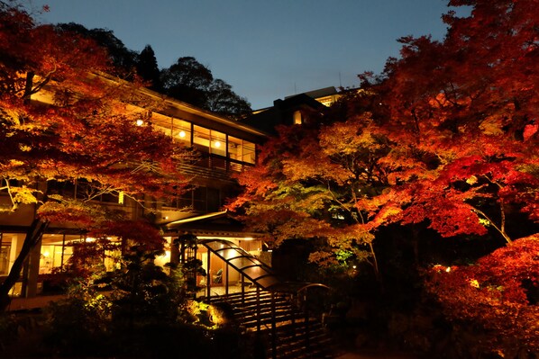 Une maison traditionnelle japonaise avec un escalier en bois menant à l’entrée, entourée d’un feuillage automnal.