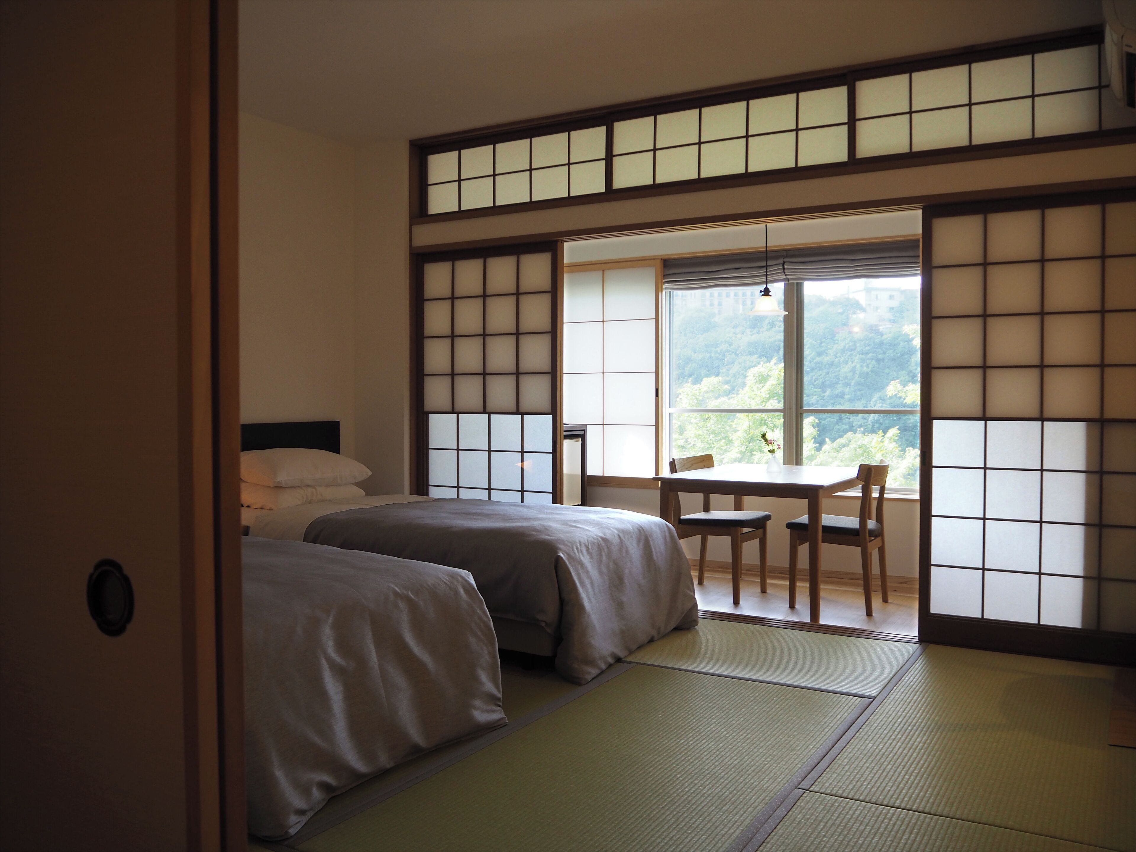 A traditional Japanese-style room with tatami flooring, a low table with chairs, and a large window offering a view of greenery.
