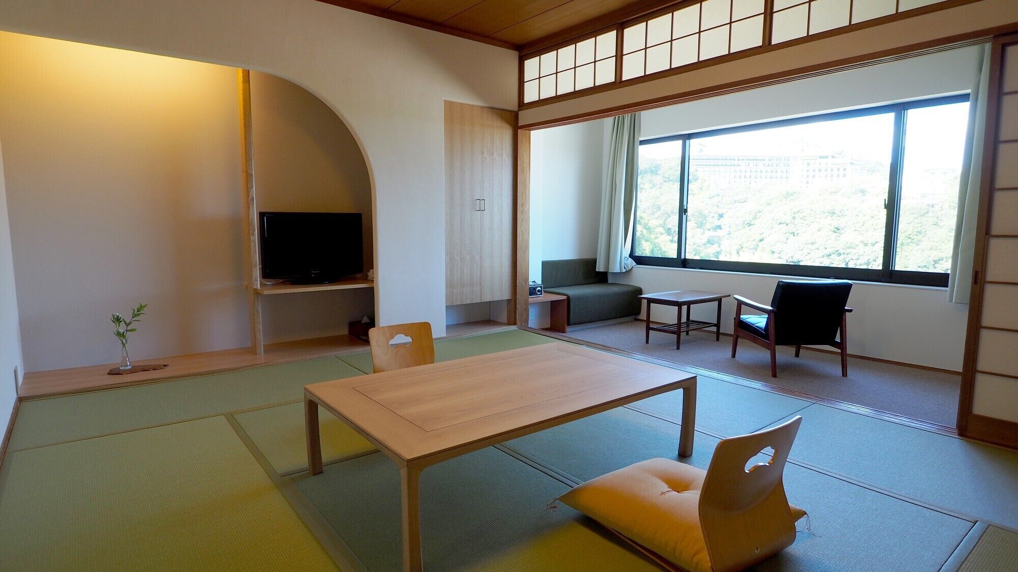 A traditional Japanese room with tatami flooring, a low table, chairs, a television, and a window with a view.