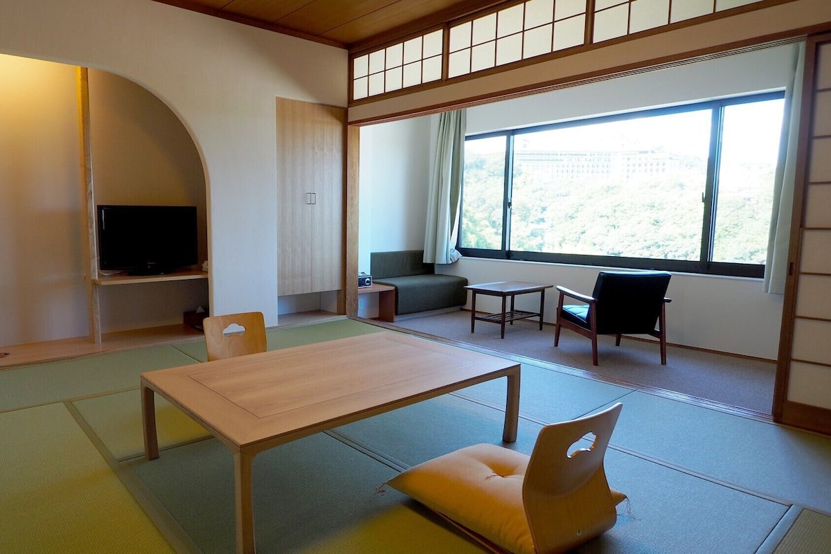 Ein traditioneller japanischer Raum mit Tatami-Boden, einem niedrigen Tisch, Stühlen, einem Fernseher und einem Fenster mit Ausblick.