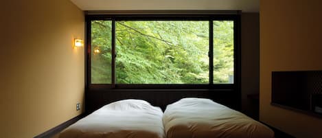Un dormitorio con un ventanal grande que da a un bosque. Hay dos camas con ropa blanca y un televisor montado en la pared.