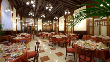 Un comedor espacioso con varias mesas redondas, cada una dispuesta con platos, vasos y cubiertos. La sala presenta paneles de madera en las paredes y el techo, y grandes ventanas con cortinas.