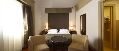 Una camera d'albergo con un letto, due comodini con lampade, un tavolino rotondo, due poltrone, un tappeto decorato e una porta che dà su un bagno.