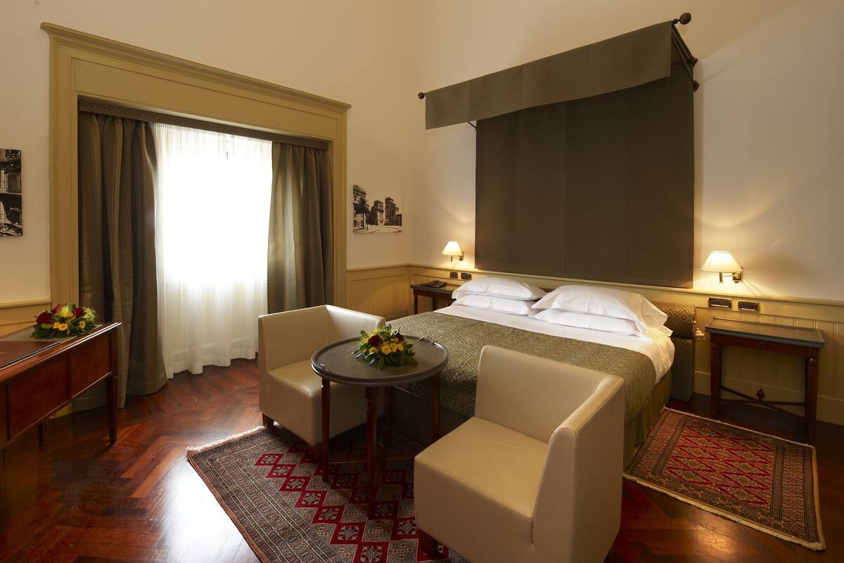 A hotel room with a bed, two armchairs, a small table with flowers, a desk with a lamp, a window with curtains, and a wall-mounted TV.