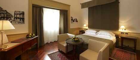 Une chambre d’hôtel comprenant un lit, deux fauteuils, une petite table avec des fleurs, un bureau avec une lampe, une fenêtre avec des rideaux et un téléviseur fixé au mur.