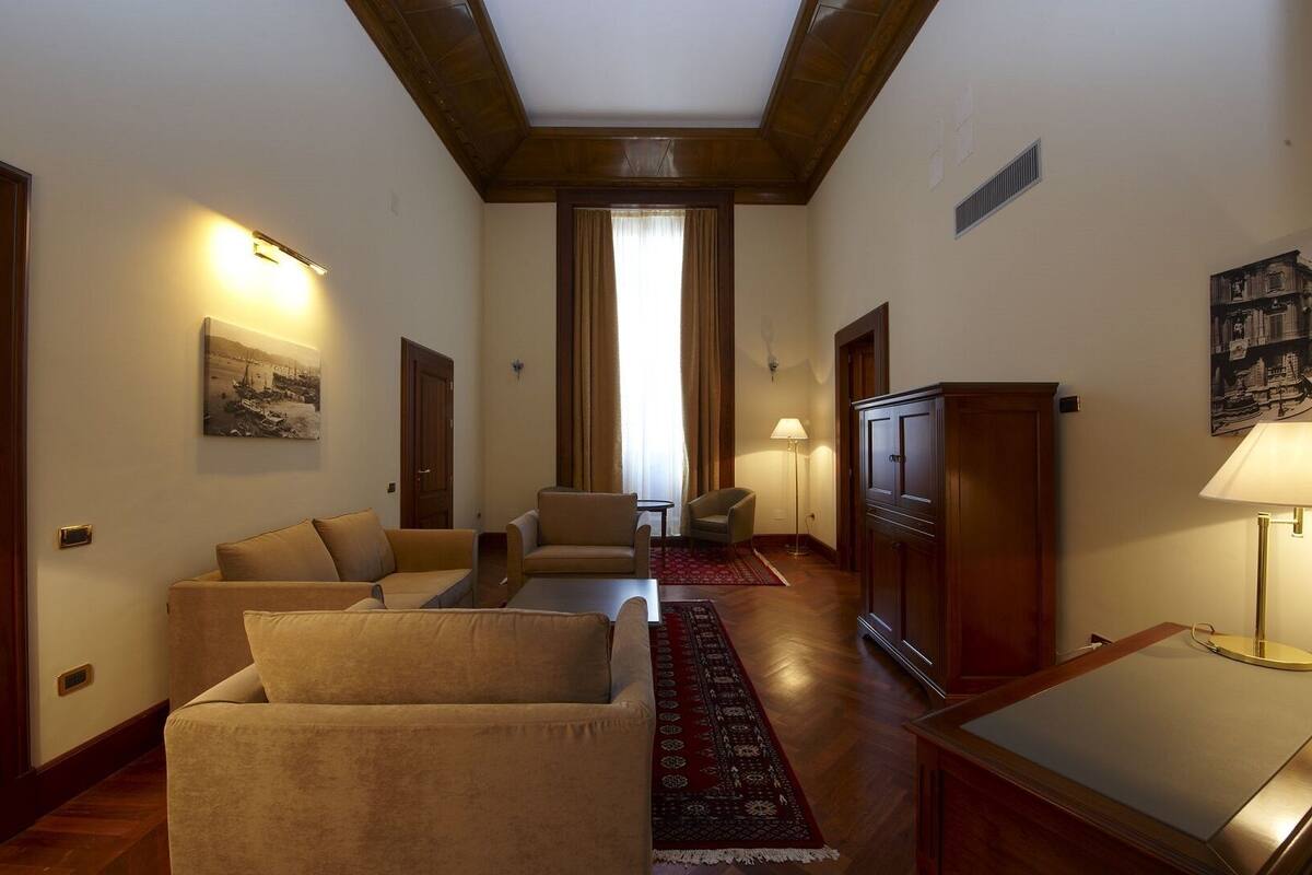 A spacious living room with a high ceiling, wooden beams, a large window with curtains, a red rug, and a cabinet.