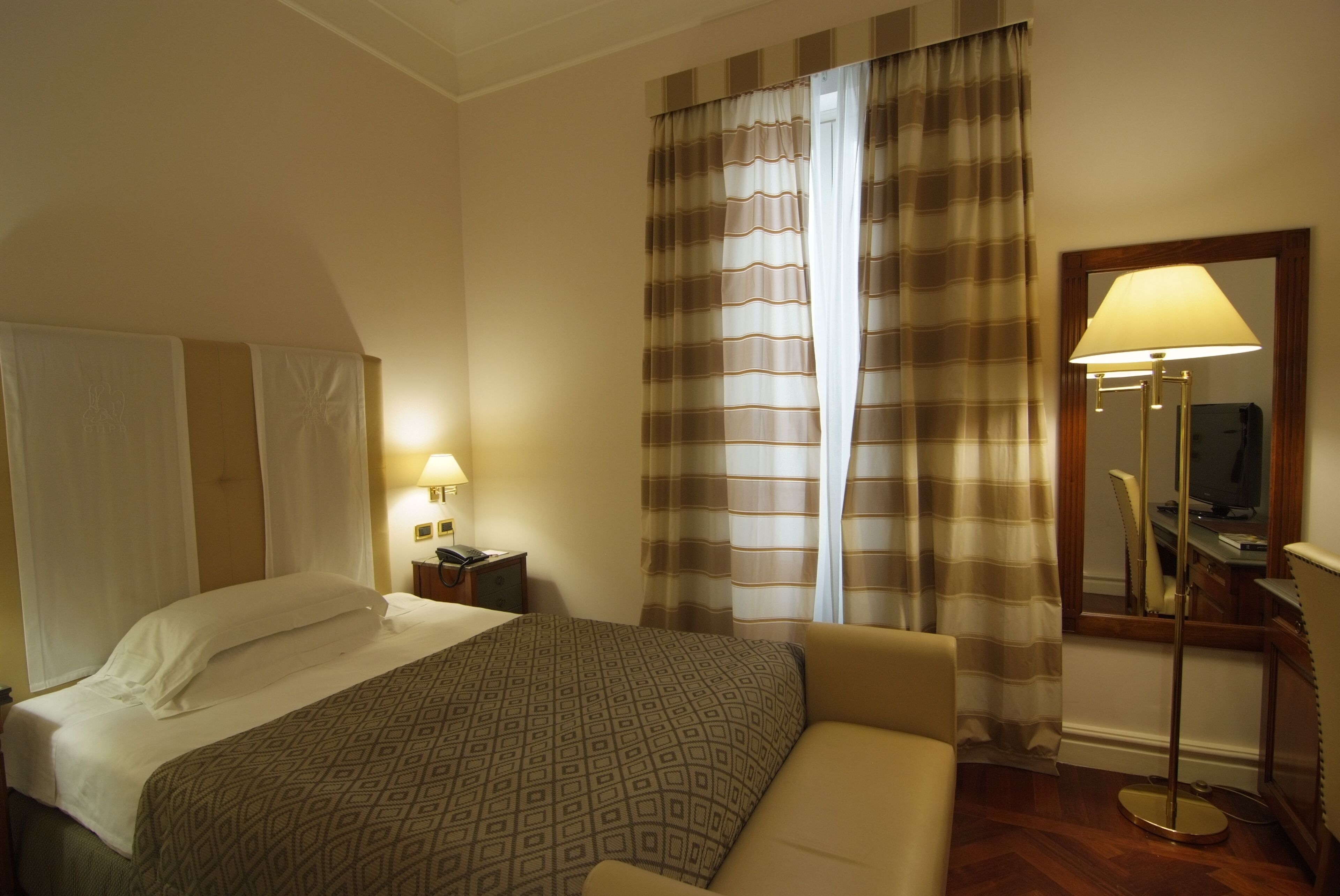 A hotel room with a bed, a bedside table with a lamp, a chair, a desk with a computer, and a large mirror.