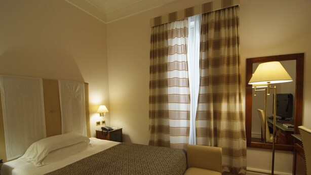 Habitación de hotel con cama, mesita de noche con lámpara, una silla, escritorio con ordenador y un espejo grande.
