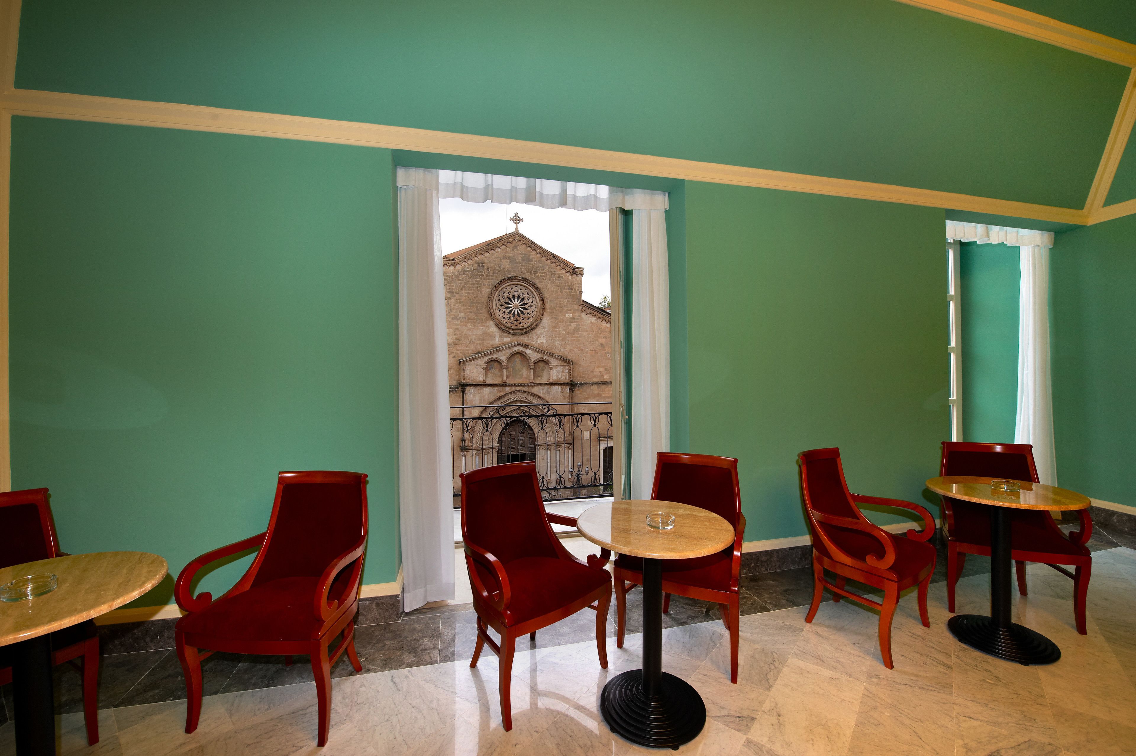 Un espace repas avec des chaises rouges et des tables rondes, une vue sur une église par la fenêtre et des murs verts.
