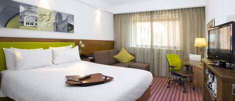 A hotel room with a bed, a desk, a chair, a television, and artwork on the wall.