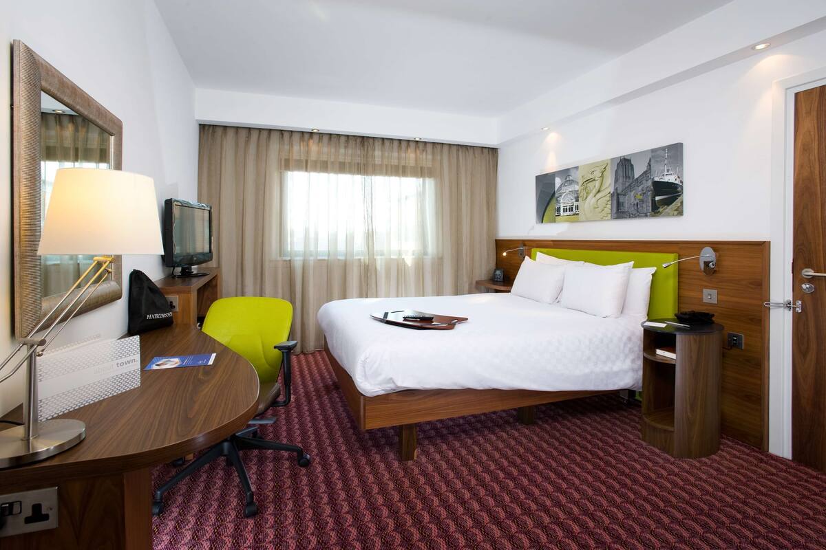 A hotel room with a bed, a desk with a lamp, a chair, a TV, and a painting on the wall.