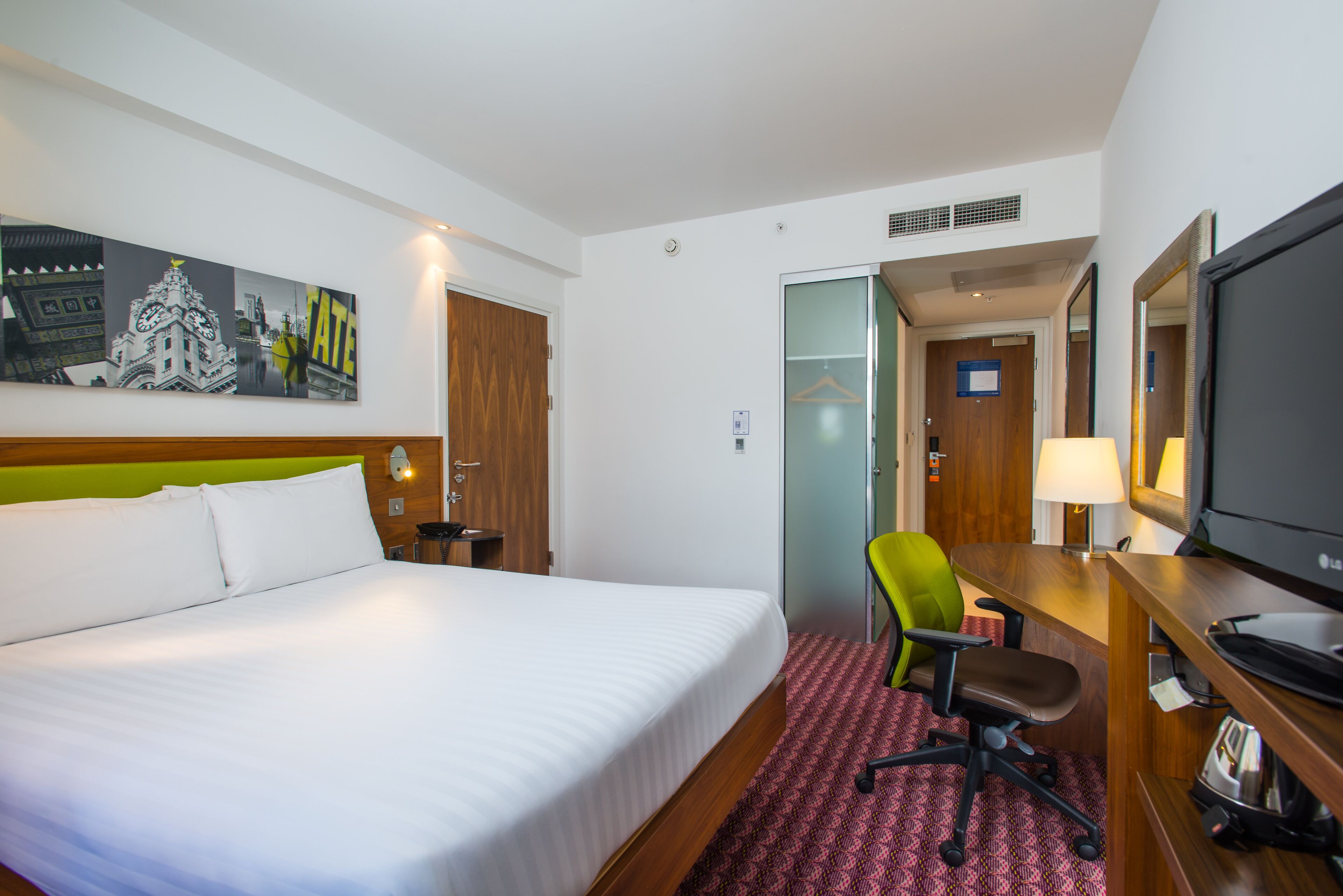 A hotel room with a large bed, a desk with a computer, a chair, a TV, and a painting on the wall.