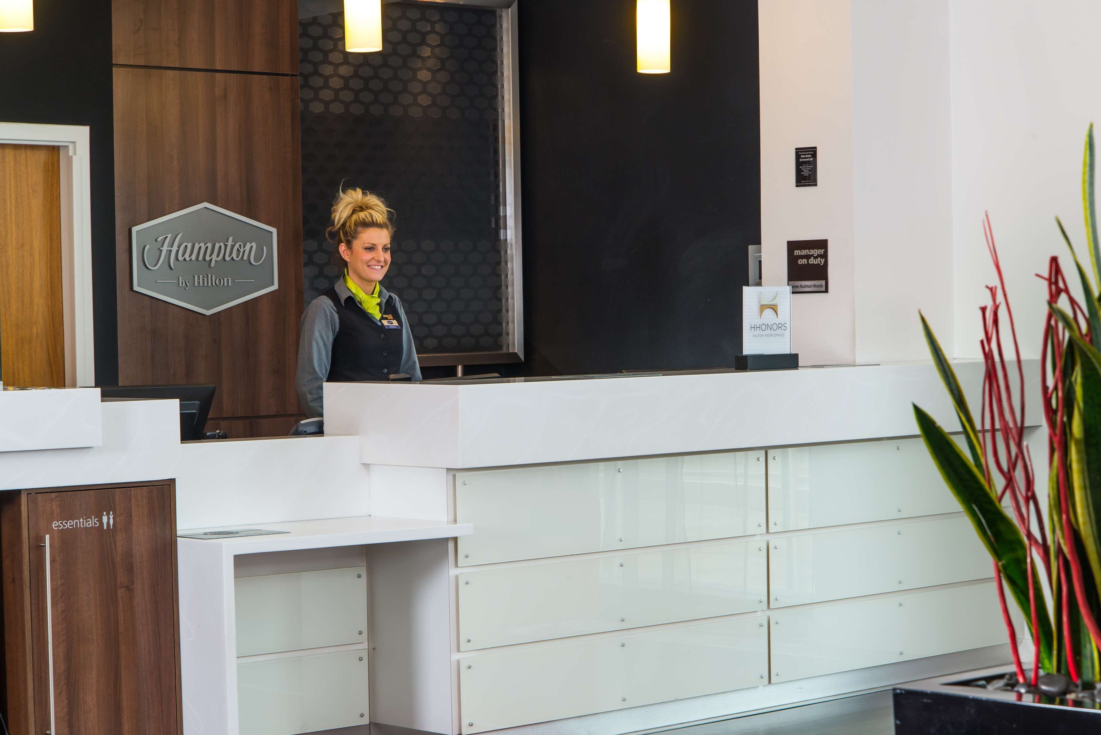 A reception desk at Hampton by Hilton with a modern design, a sign indicating the location, and a manager on duty sign.