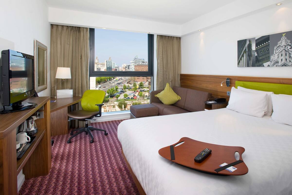 A hotel room with a bed, a desk with a computer, a chair, a TV, and a window with a city view.
