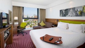 Habitación de hotel con una cama, un escritorio con computadora, una silla, un televisor y una ventana con vista a la ciudad.