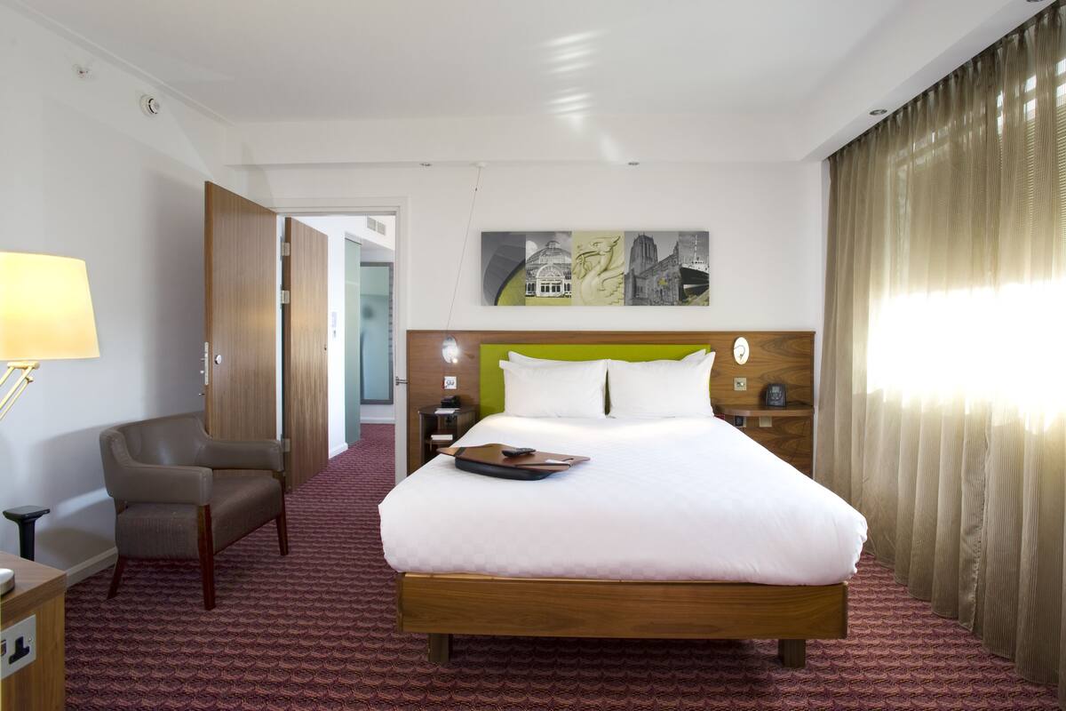 A hotel room with a large bed, a desk with a lamp, a chair, and a painting on the wall.