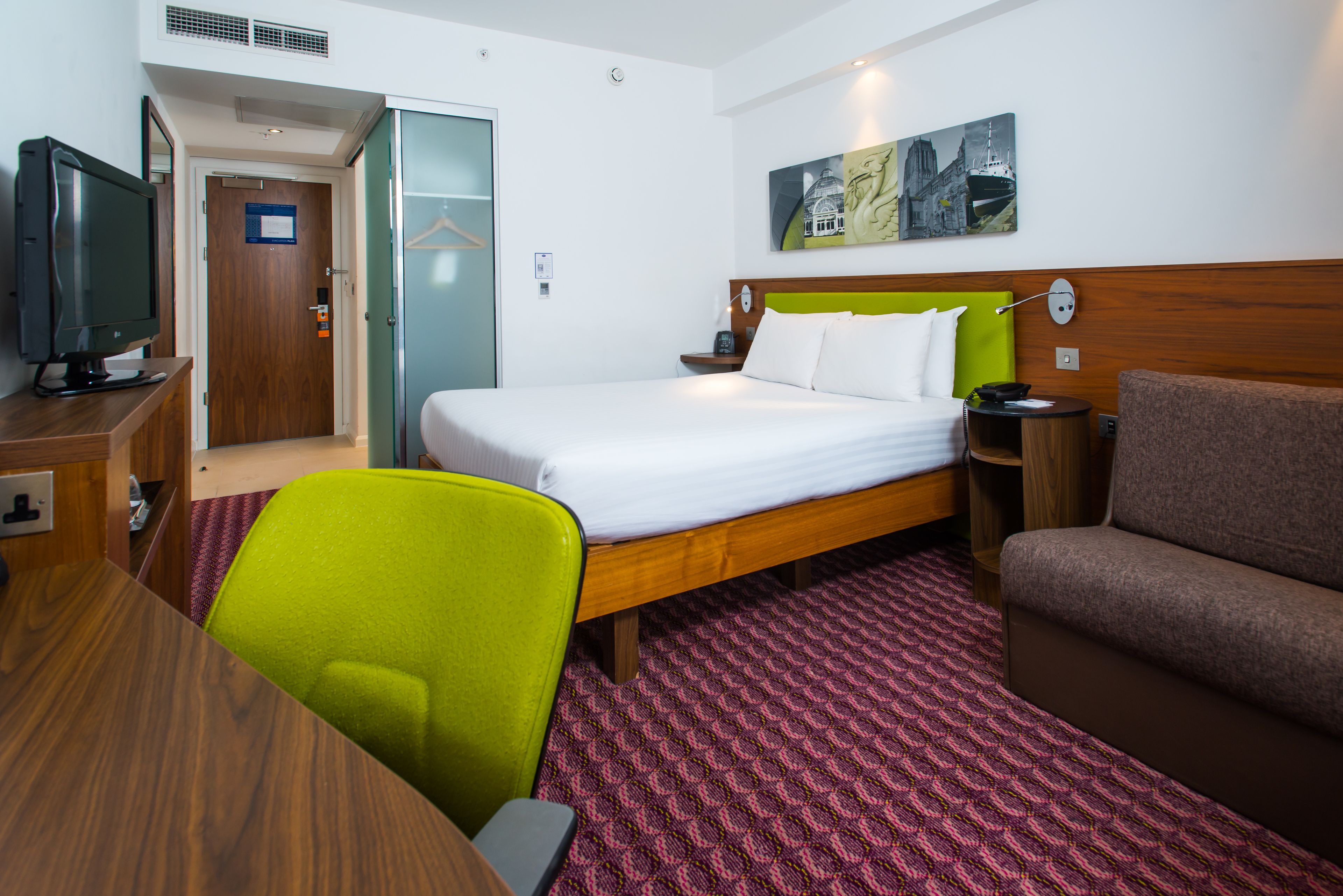 Habitación de hotel con cama, escritorio con televisor, silla verde, sofá marrón y alfombra estampada.