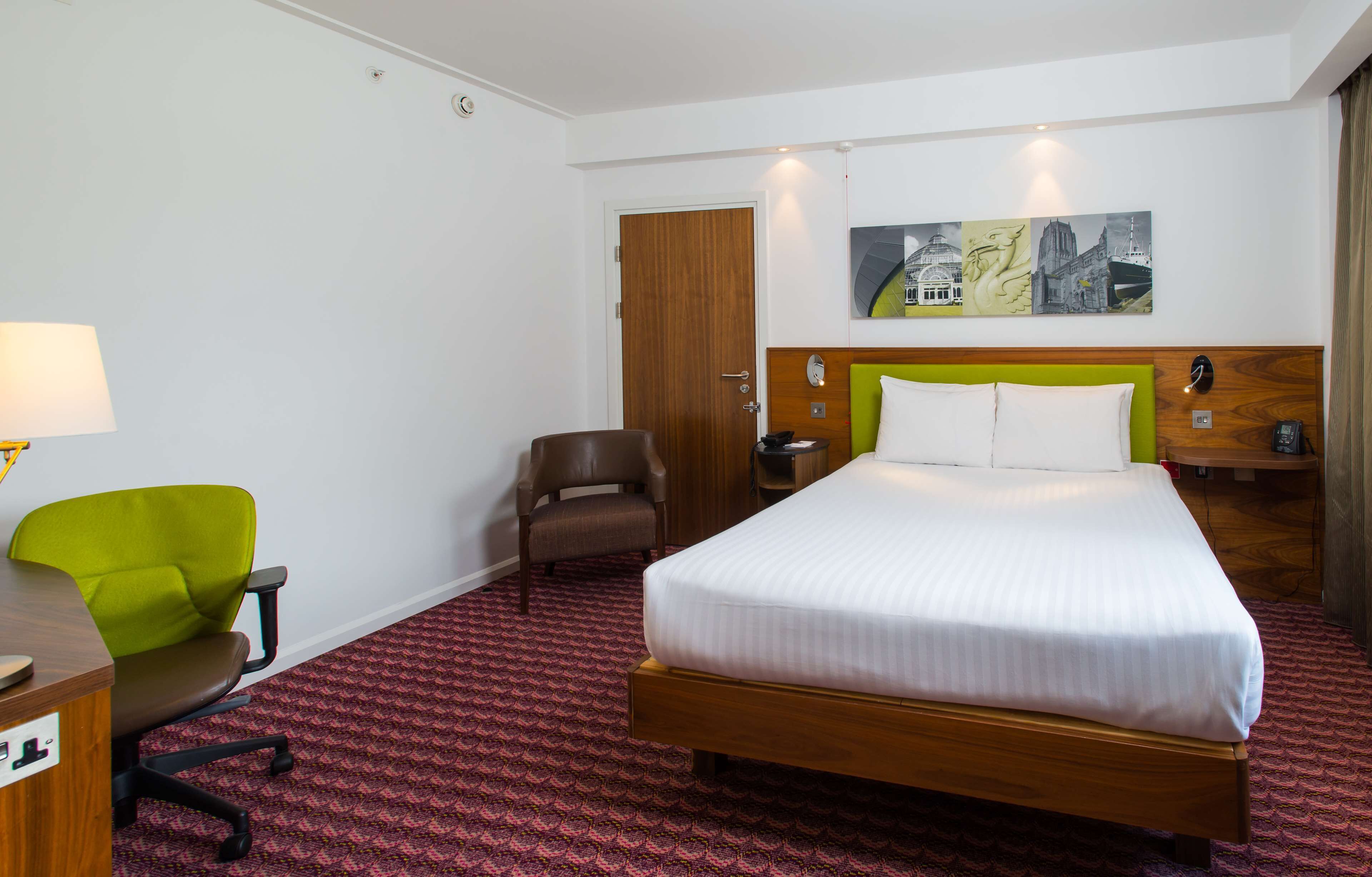 Habitación de hotel con cama, escritorio con lámpara, silla verde, sillón marrón y un cuadro en la pared.