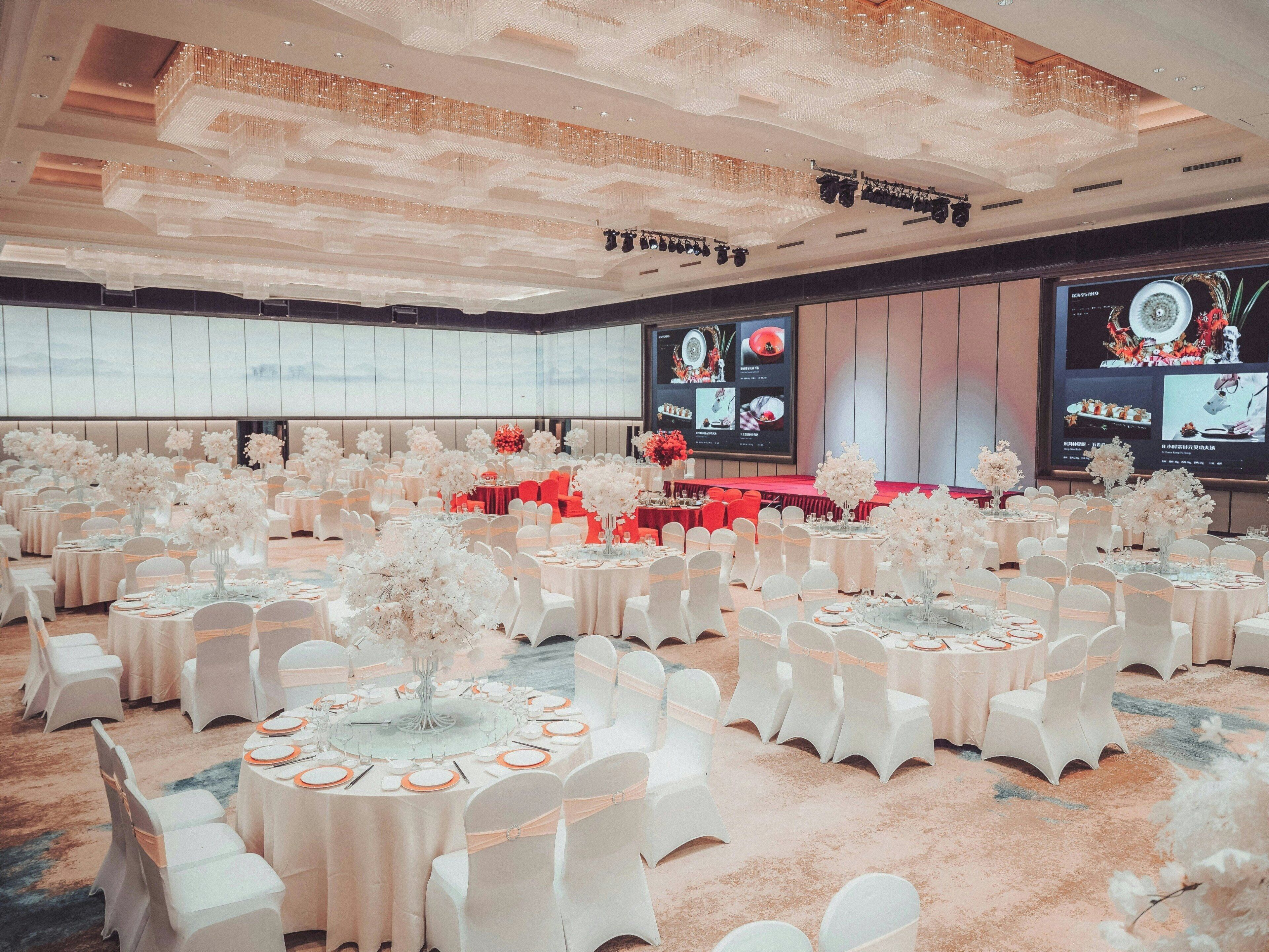 Een feestzaal met ronde tafels, witte stoelen en een podium met een rode achtergrond.