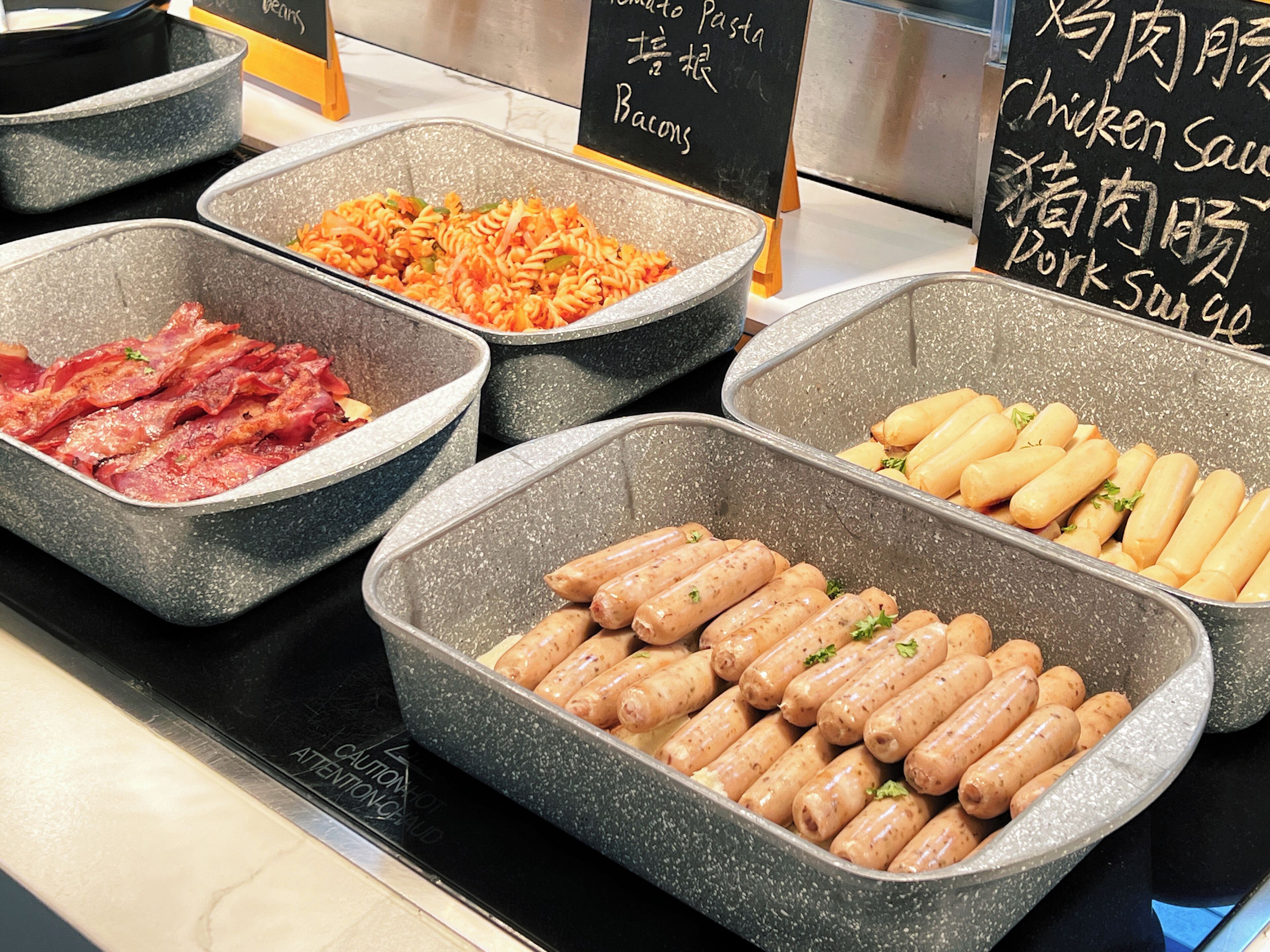 Een buffet met diverse gerechten, waaronder spek, pasta, worst en verschillende sauzen.