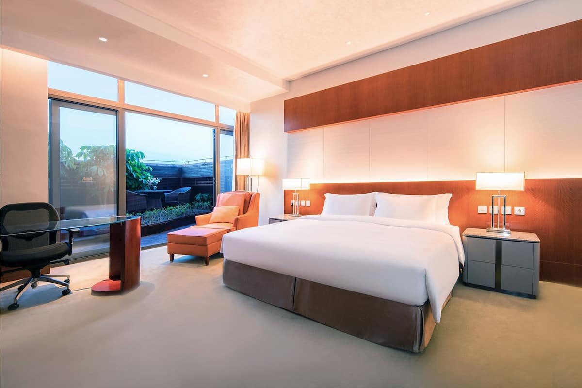 Ein modernes Hotelzimmer mit einem großen Bett, zwei Nachttischen inklusive Lampen, einem Schreibtisch mit Stuhl und Blick ins Grüne.