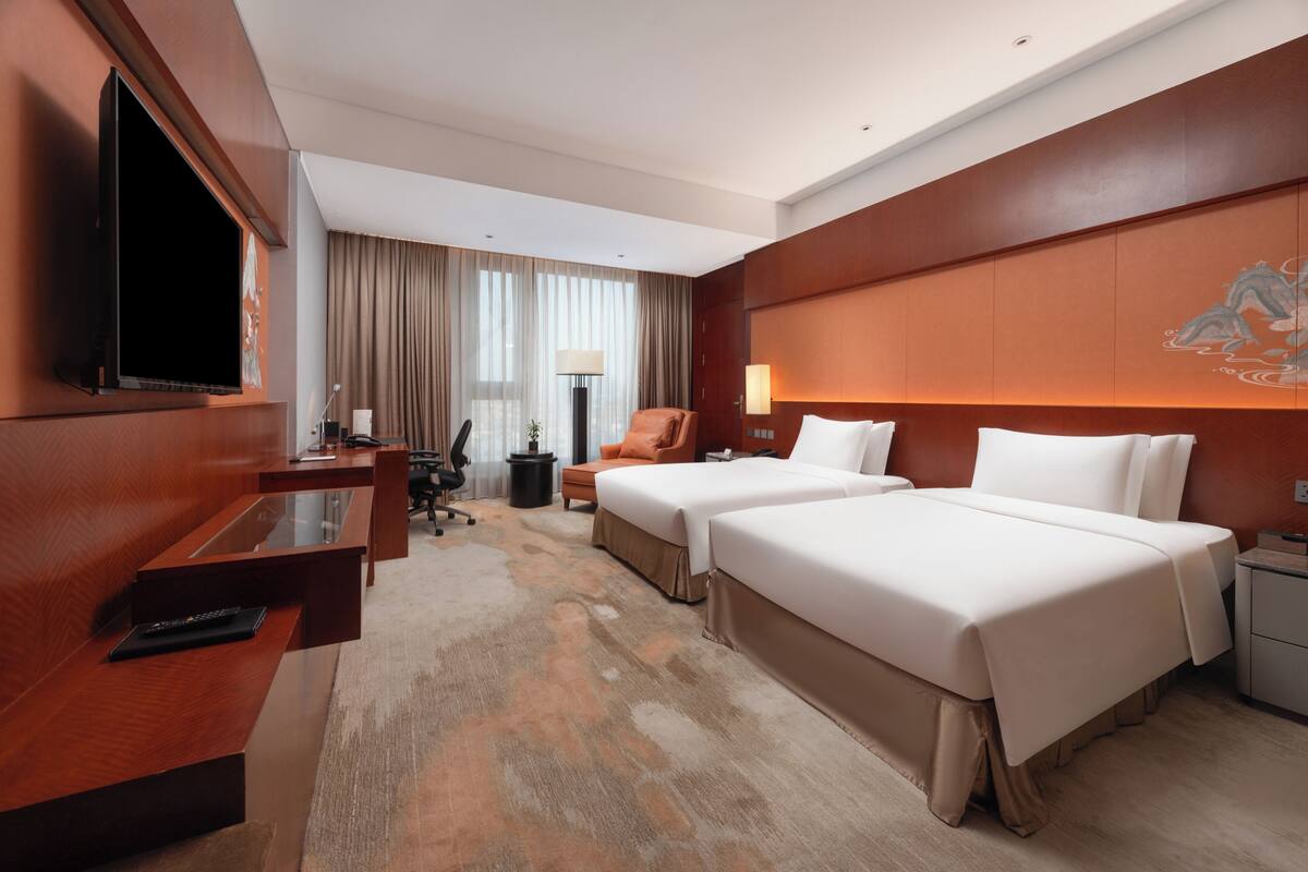 A hotel room with a bed, two bedside tables, a desk with a flat-screen TV, a chair, a lamp, and a large window with curtains.