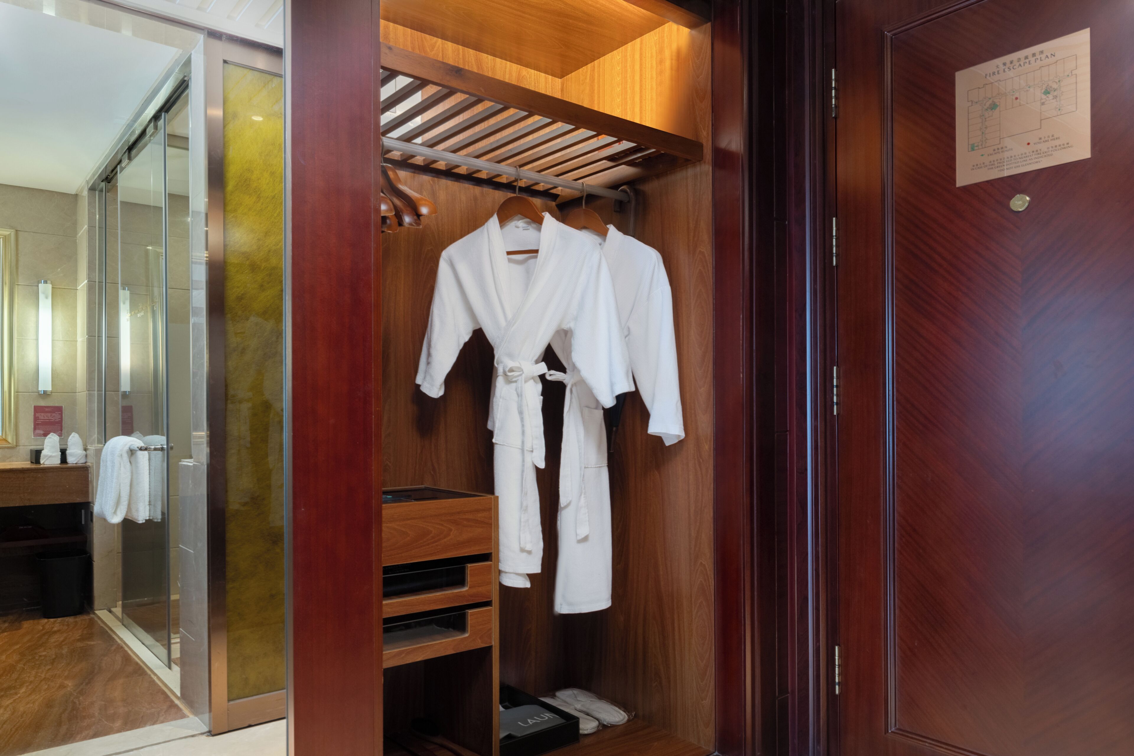 Un closet espacioso de madera con dos batas blancas colgadas en un perchero, un estante de madera con zapatos, y una puerta con una placa.