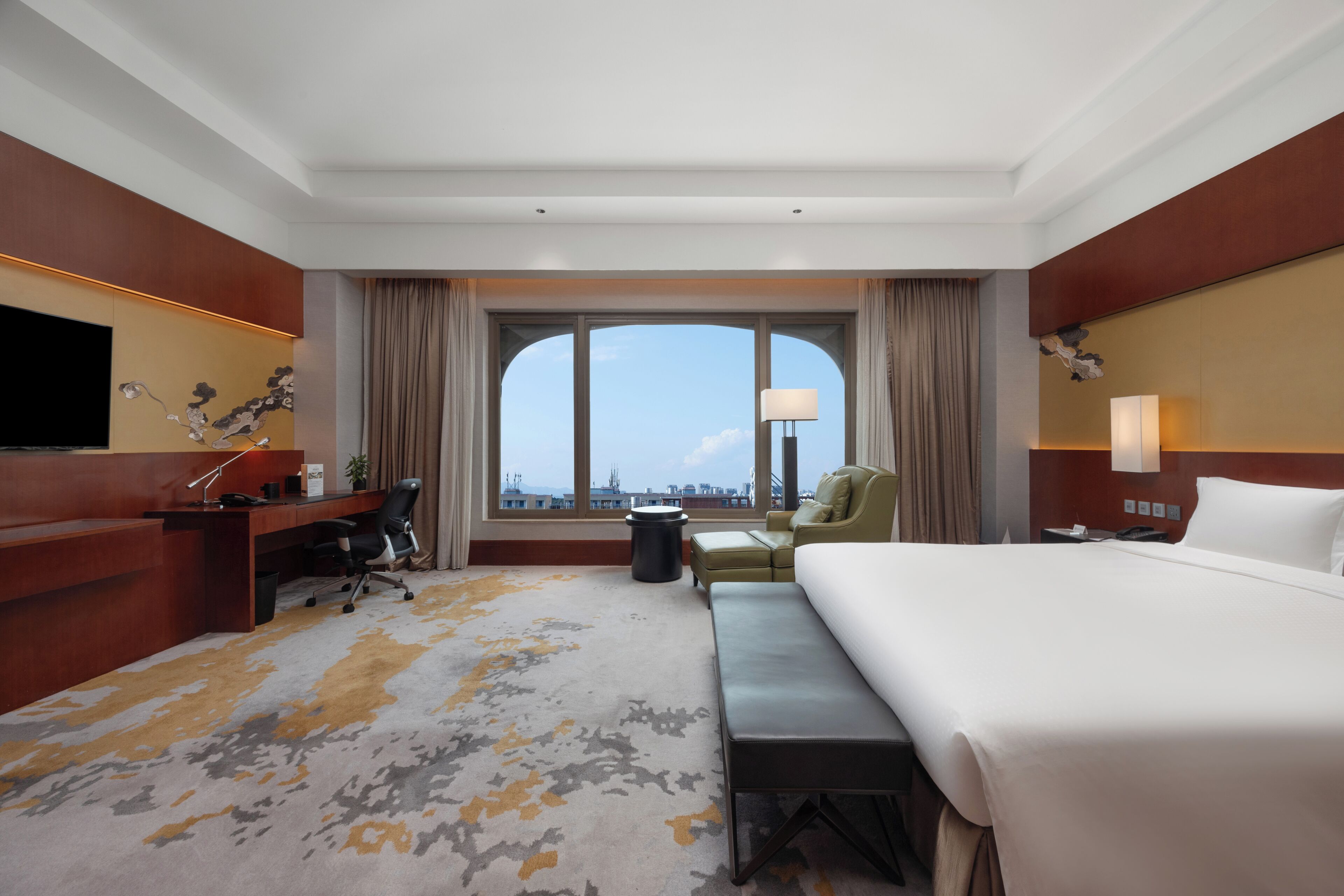 A hotel room with a large bed, a desk with a chair, a sofa, a TV, and a window with a view of buildings.