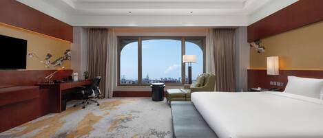 A hotel room with a large bed, a desk with a chair, a sofa, a TV, and a window with a view of buildings.