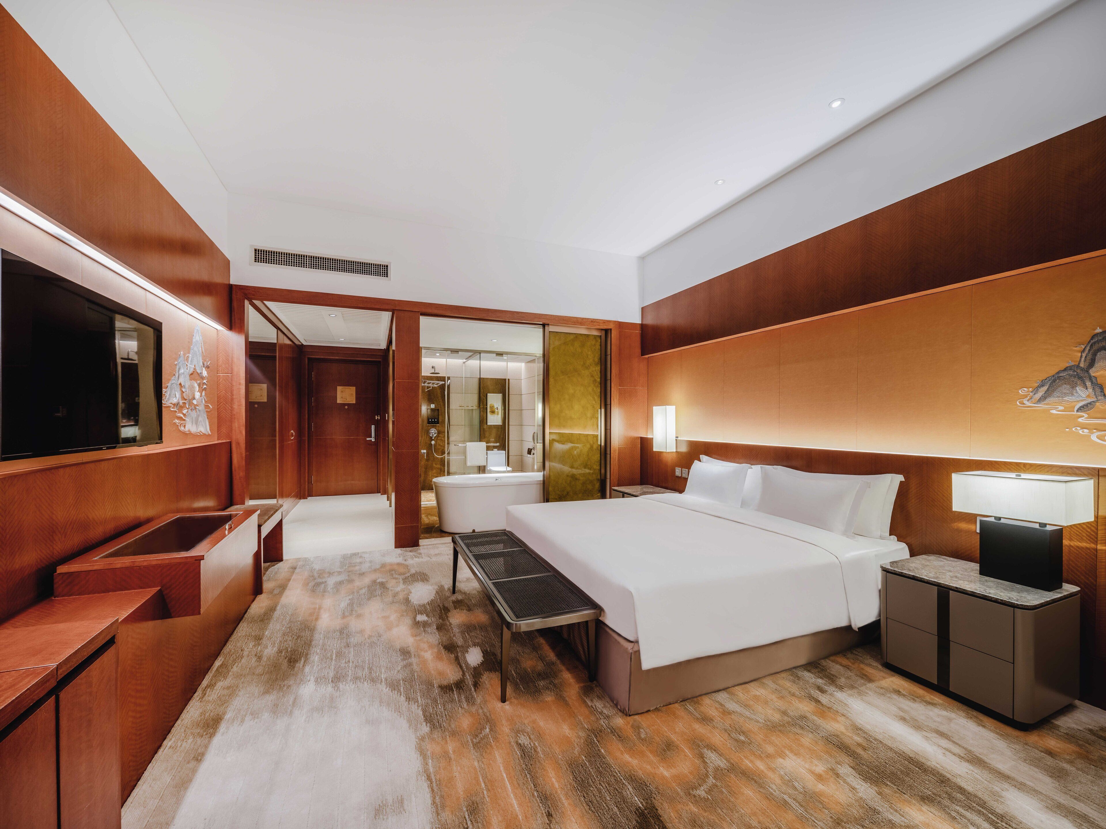 A modern hotel room with a large bed, a desk, a TV, and a bathroom visible through an open door.