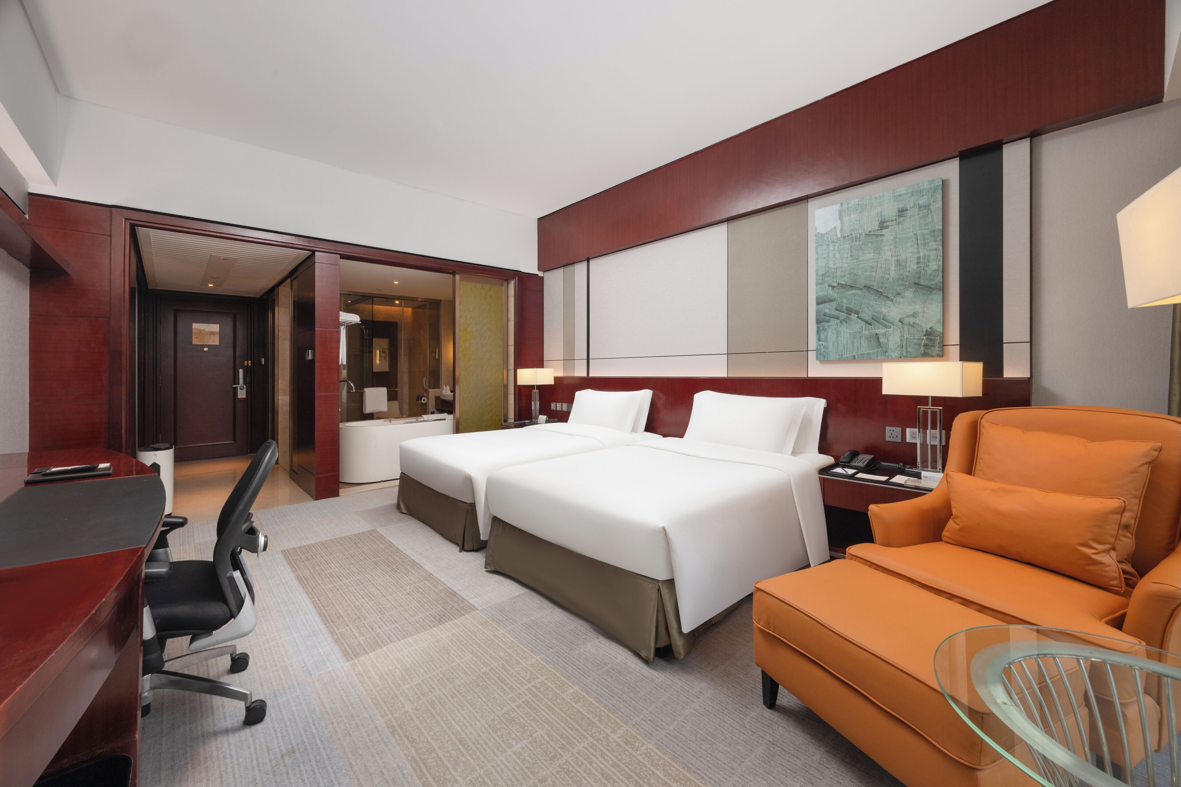 A hotel room with a bed, two bedside tables, a desk, a chair, an orange sofa, and a glass coffee table.