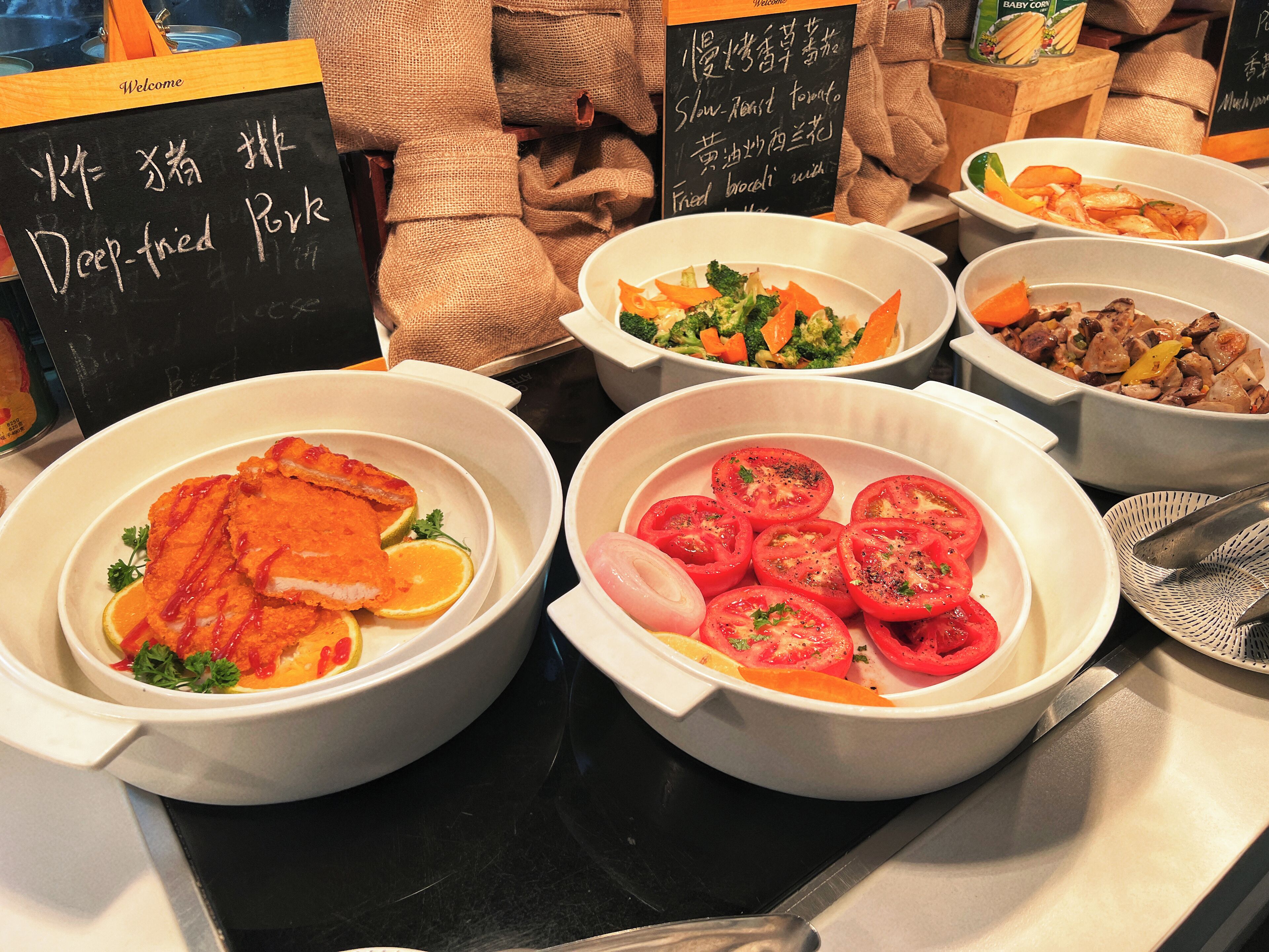 Un buffet proposant divers plats, notamment du porc frit, des brocolis sautés et des tranches de tomates.