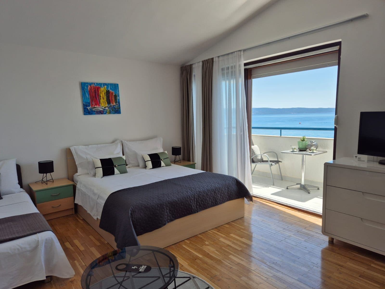 A hotel room with a bed, a desk, a chair, a TV, and a balcony with a view of the sea.