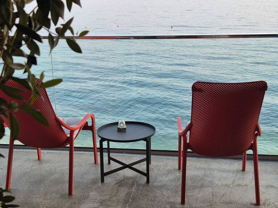 Two red chairs and a small table on a balcony with a view of the sea.
