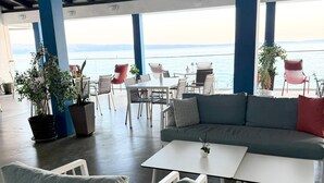 Un espacio al aire libre con techo, mesas y sillas, plantas en macetas y vista al mar.