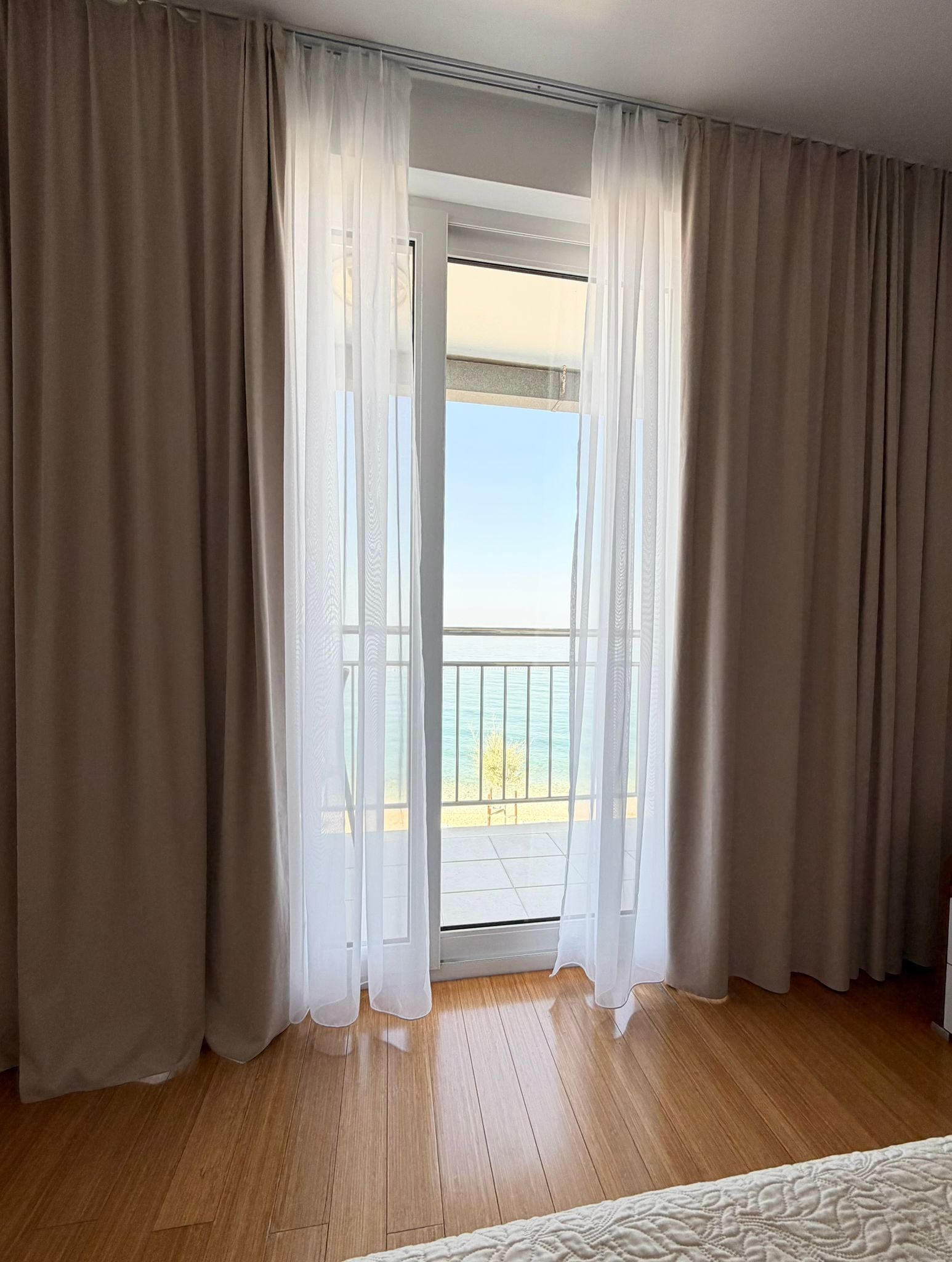 Una habitación con piso de madera, un balcón con vista al mar y cortinas color crema.