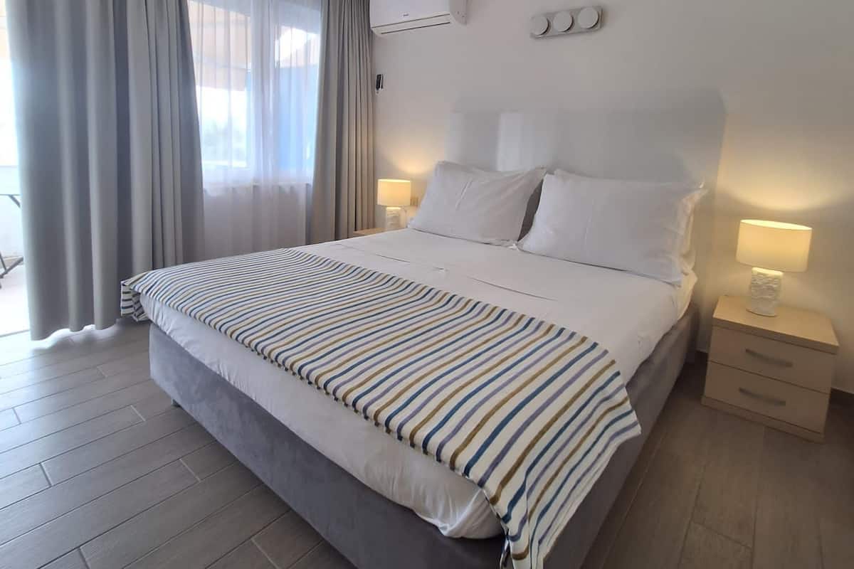 Habitación de hotel con una cama grande, una mesita de noche con lámpara, un equipo de aire acondicionado y una ventana con cortinas.
