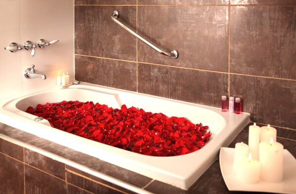 A bathtub filled with red rose petals, a grab bar, a white sink with candles, and toiletries. - Be Hollywood Hotel (Buenos Aires)