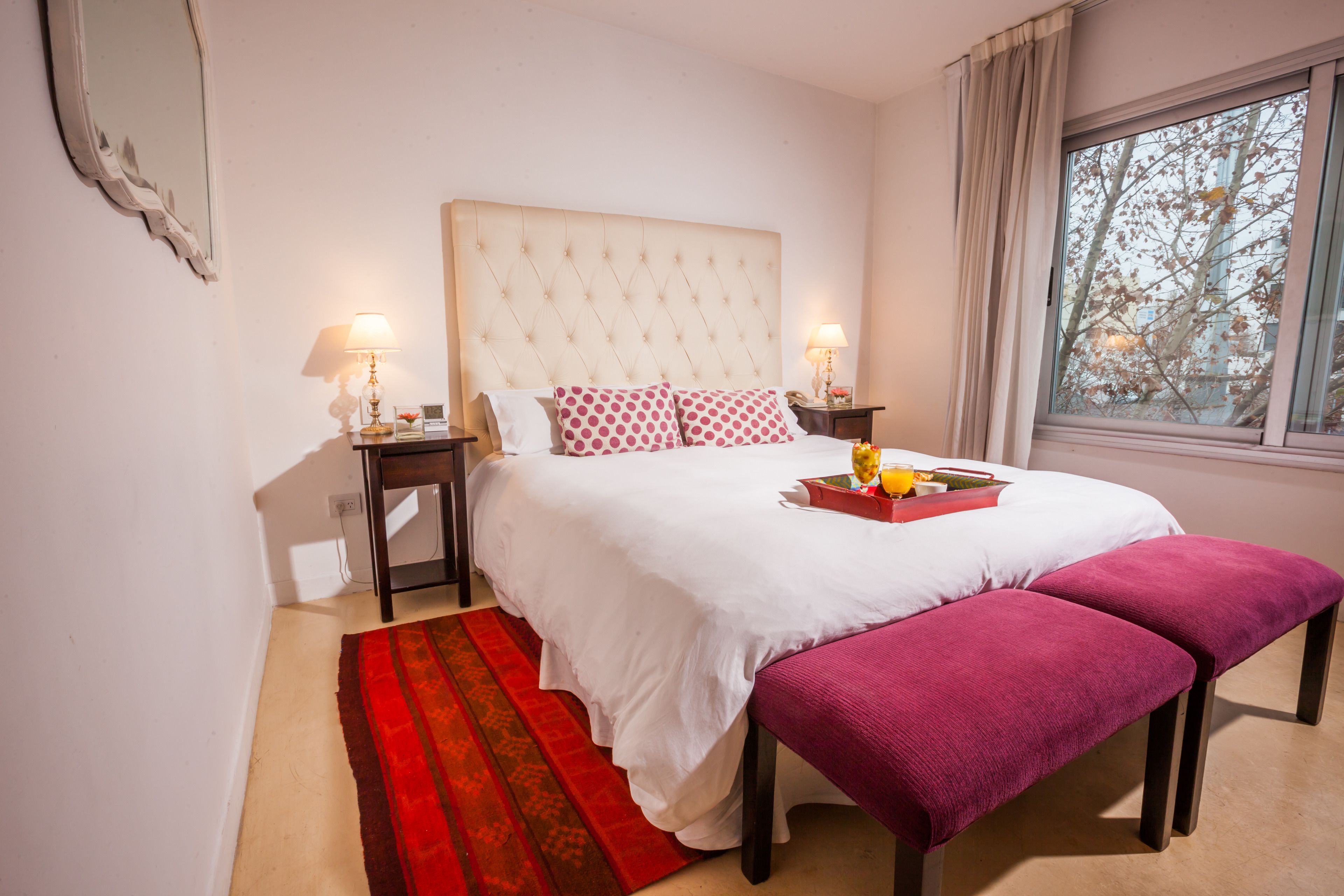 A bedroom with a large bed, two bedside tables, a window with a view, and a red and white patterned rug.